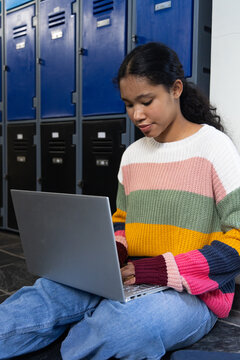 Female child sitting on tiled floor by school lockers holding silver laptop wearing striped sweater