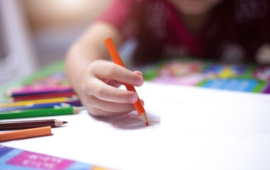 Child draws with red crayon on white paper. Hands grip crayon, showing focus and care. Bright crayons lie nearby, colorful and unused. Soft background blurs, drawing attention to action © Issarawat Tattong