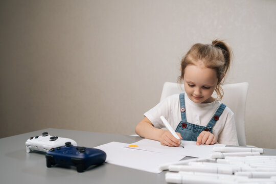 Focused young girl in overalls sitting at table drawing with markers. Kid ignores video game controllers, choosing creative hobby over digital entertainment. Soft light, neutral colors, copy space.