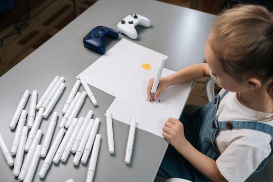 High angle view of little girl focused on drawing with markers on white paper. Many pens on grey desk next to video game controllers. Concept of creative childhood and hobbies.