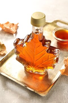 Leaf shaped bottle of maple syrup, bowl and dried leaves on light grey table, closeup