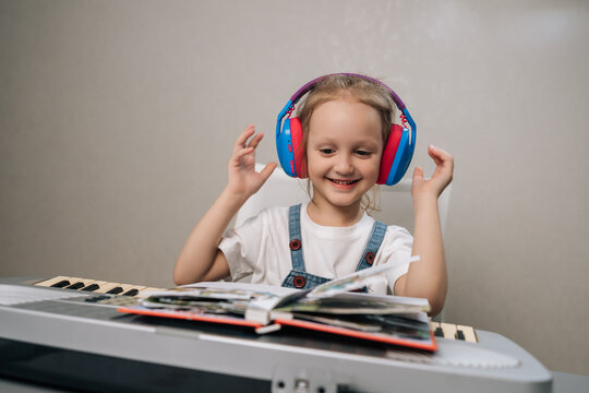 Joyful young student in colorful headphones smiling while taking online music lesson. Girl sitting at electronic keyboard, learning from book. Concept for homeschooling, e-learning, childhood hobbies.