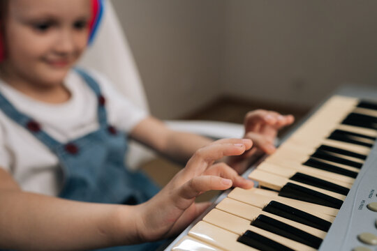 Selective focus on little girls hands playing digital piano. Blurred smiling child in headphones learns music. Concept of childhood education, hobby, creative development at home.