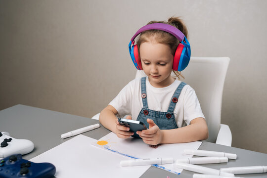 Portrait of young focused girl in headphones sitting at table using mobile phone. Modern childhood concept with technology, gaming controllers, creative drawing tools for education and entertainment.