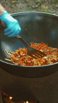 A person wearing blue gloves is stirring food in a black pot outdoors, possibly in a camping or cookingA.