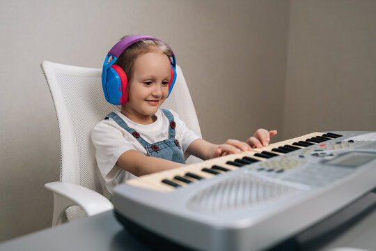 Young girl wearing colorful headphones practices keyboard synth at home, smiling and focused while developing musical skills, creativity and joyful childhood learning.