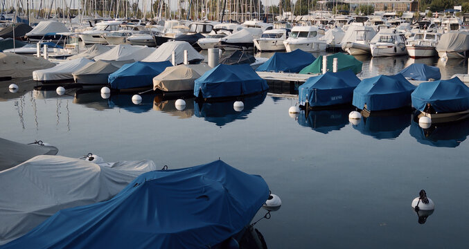 Covered with tarps, the yachts in the Geneva marina line the pier in soft light, their tall masts rising over the still lake.