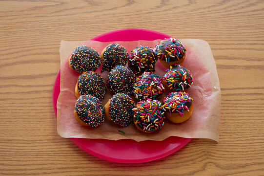 A dozen donut holes with rich, chocolate icing and colorful sprinkles sitting on a sheet of wax paper on a plate.