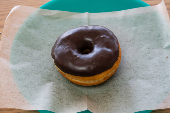 A round donut with rich, chocolate icing sitting on a sheet of wax paper on a plate.