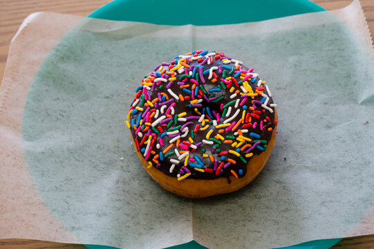 A round donut with rich, chocolate icing and colorful sprinkles sitting on a sheet of wax paper on a plate.
