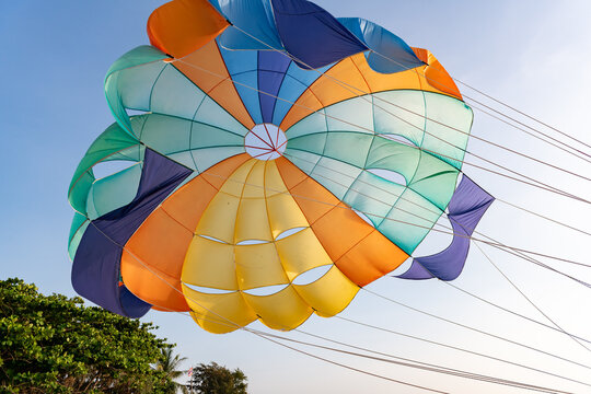 Vibrant multi-colored parachute canopy filled with wind, soaring against a clear blue sky, with green foliage visible at the bottom, invoking summer fun and adventure.