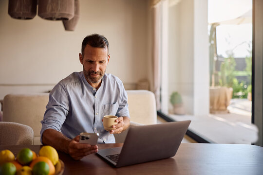Smiling freelancer using cell phone while drinking coffee and working on laptop at home.