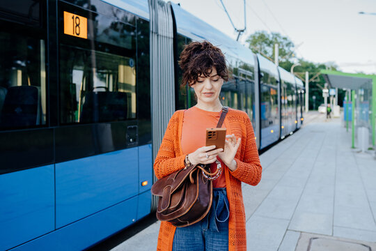 Connected young woman checking transit app while waiting beside city tram