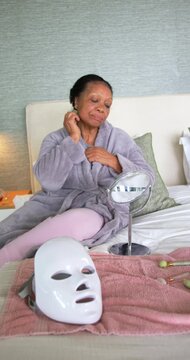 Vertical video: Senior woman starting facial rolling jade roller and checking mirror on bed in robe