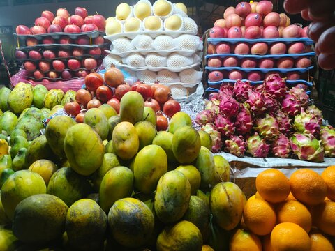 Street market display featuring stacked yellow and red apples mangoes dragon fruits and fresh oranges piled neatly for customers to purchase during the day at a food bazaar stall.