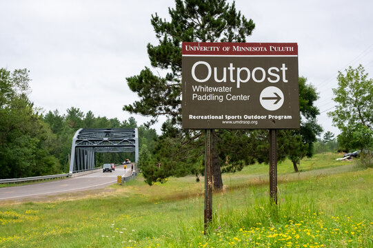 CARLTON, MINNESOTA &mdash; JULY 25, 2026: Sign for the University of Minnesota Duluth (UMD) "Outpost" Whitewater Paddling Center with a truss bridge and cars in the distance.