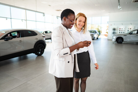 Two diverse women looking at a smartphone screen in a modern car dealership showroom