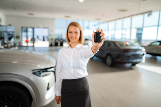 Young adult woman sales consultant holding car key in auto dealership showroom, smiling