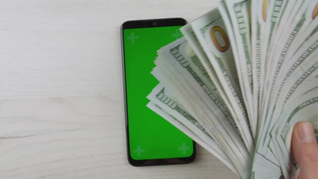 A mobile phone with a blank green display is placed beside a stack of US dollars on a clean white surface. This image represents digital finance, cashless payments, and mobile banking trends