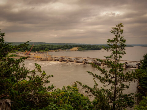 Distant view of a large river lock and dam system surrounded by lush green forests under an overcast cloudy sky