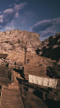 Wooden structures and an empty wagon are located in an old mining area. The setting has rocky hills and clear blue skies. The sun shines on the scene during the day.