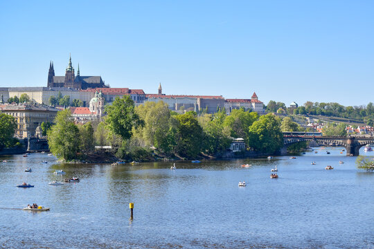 A panoramic view of Prague from the Vltava River, featuring Prague Castle and St. Vitus Cathedral above the historic city, with Charles Bridge and boats visible in the foreground