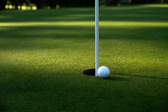 White golf ball tap in close to hole and pin on putting green, early morning sunny summer day on the golf course, relaxing recreational sport

