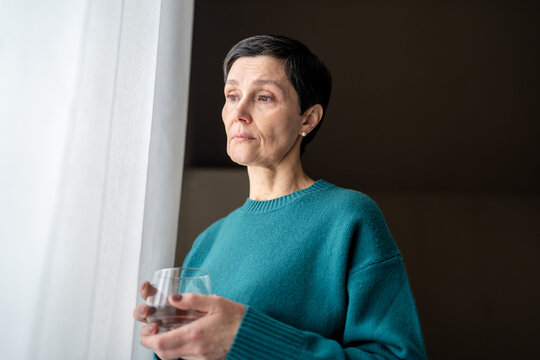 Depressed mature woman with water glass waiting at window lost in thought. Nervous tension, self soothing, deep contemplation loneliness, emotional pause, private dilemma, silent endurance