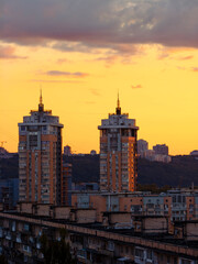 Fototapeta premium Kyiv's modern residential high-rises are standing prominently against a vibrant orange and yellow sunset sky, illuminating the urban landscape with warm evening light as clouds gather overhead