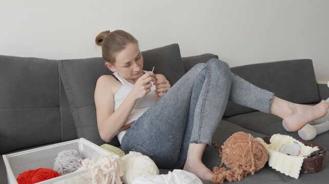 A relaxed woman enjoys knitting on a stylish couch, surrounded by various yarns and knitting supplies. The scene captures a peaceful moment of creativity and mindful crafting at home