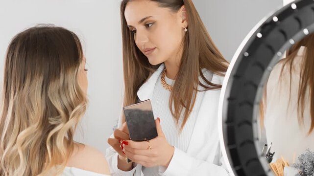 A beauty expert carefully works on a client&rsquo;s makeup, focusing on detail and precision. The scene captures a trendy, professional environment with natural lighting