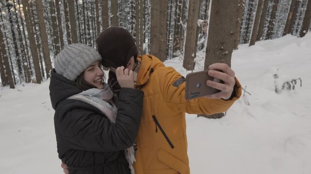 Happy couple embraces and smiles while taking a selfie in a snow covered forest, surrounded by tall trees. Their winter clothing and cheerful expressions highlight a cozy outdoor adventure