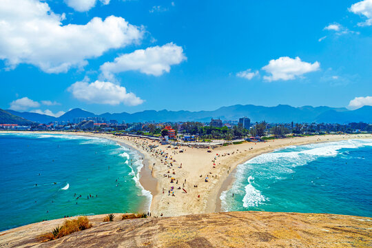 Stunning aerial view of the sandy strip connecting Macumba and Recreio beaches in Rio de Janeiro, Brazil, featuring vibrant blue ocean waters, mountains, and tourists enjoying a sunny day.