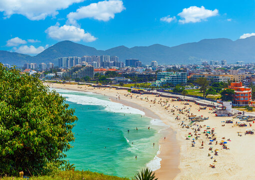 Aerial view of Praia da Macumba, Rio de Janeiro. Vibrant sunny day featuring turquoise ocean waves, a crowded sandy beach, coastal cityscape, and scenic mountains in the background.
