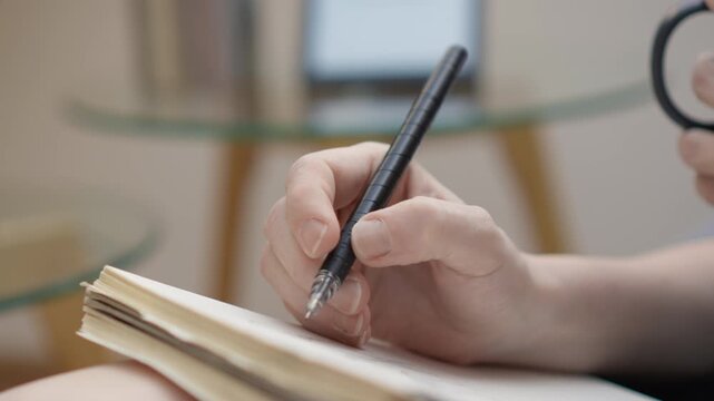 A hand holds a black pen and writes in an open notebook, capturing the act of note taking or journaling. The blurred background suggests a calm, focused atmosphere for creativity