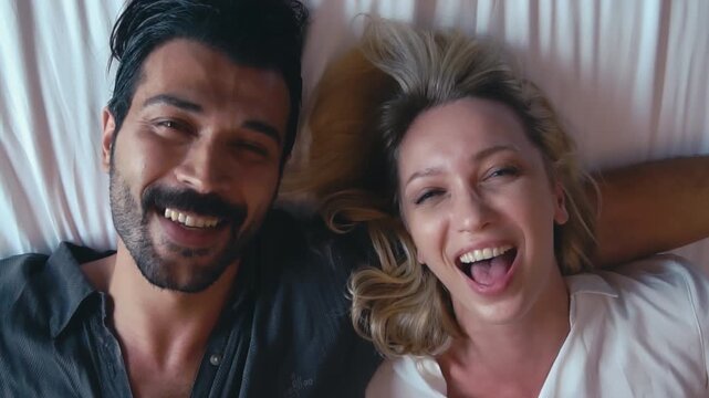 Happy young Caucasian couple lying together in bed looking at camera with big smiles and laughter. Top view portrait romantic loving husband and wife sharing intimacy and cheerful moments in bedroom