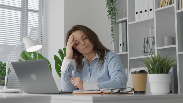 Exhausted female office worker experiencing stress and burnout while working on laptop at bright contemporary desk. Concept of overwork, mental fatigue, and work life balance in corporate environment