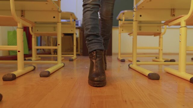Person in fashionable boots, jeans strides purposefully between rows empty desks in brightly lit classroom. Scene captures sense movement, confidence, contemporary school atmosphere