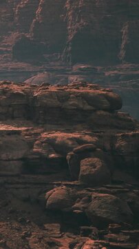 Rock formations rise in a desert area under bright sunlight. Shadows stretch across the ground as the land shows signs of erosion. The scene displays rocky textures and layered geology.