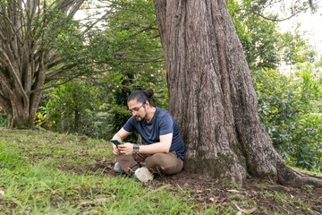 Fototapeta premium Man using smartphone sitting under tree trunk in green park