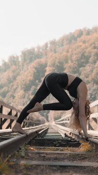 Young woman in athletic wear does yoga on abandoned railway tracks, surrounded by golden autumn trees. The serene outdoor setting emphasizes balance, mindfulness, and healthy lifestyle