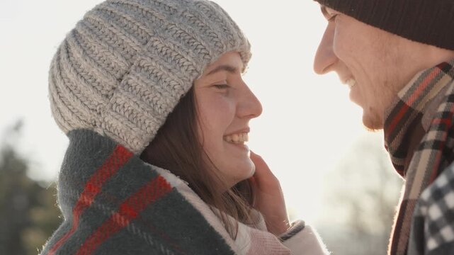 A happy young couple dressed in knitted hats and scarves share a tender moment outside in winter. Their faces are close, wrapped in a blanket, radiating warmth and affection together