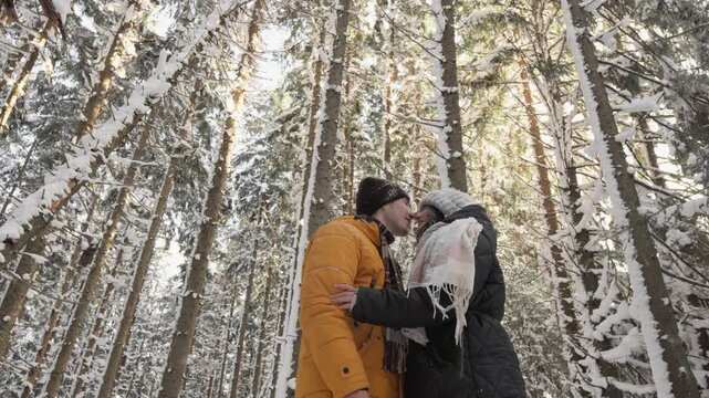 A loving couple dressed in winter clothing embraces and kisses amidst a tranquil, snow covered forest. The frosty pine trees and peaceful atmosphere create a magical, romantic winter scene