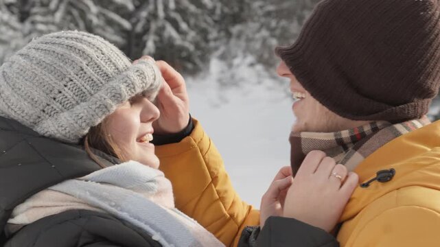 A cheerful man and woman enjoy a playful moment in a snow covered forest, dressed in winter jackets and knitted hats. Their laughter and connection create a heartwarming, festive mood