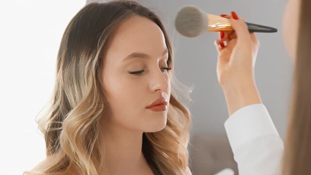 A makeup artist applies powder to a young woman's face, highlighting her flawless skin and subtle glam style. The scene captures a serene moment of beauty transformation in a contemporary setting