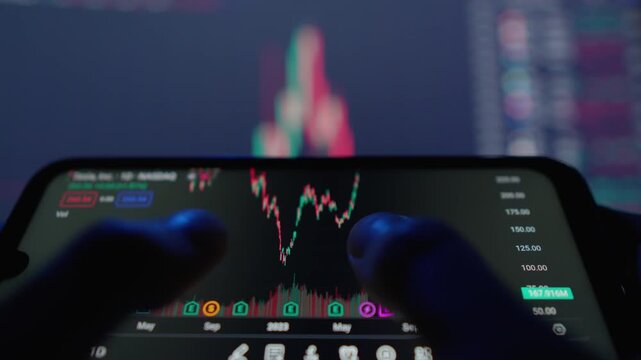 Person examines fluctuating stock market graph on mobile device, with colorful financial data displayed on screen behind. Captures essence digital trading, investment analysis