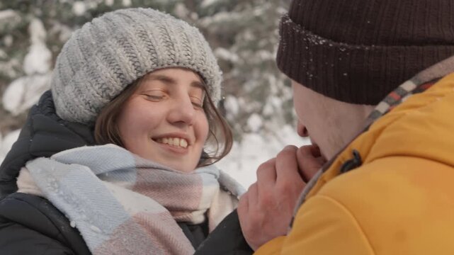 A woman in a knitted hat and scarf smiles warmly at her partner as he gently holds her hands in the snow. Their joyful connection and winter attire create a heartwarming seasonal moment