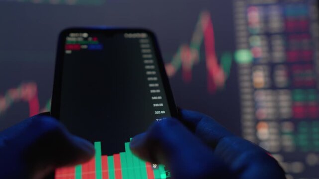 Close up hands holding smartphone showing dynamic stock market charts, with blurred monitor displaying financial graphs behind. Captures essence digital trading, market analysis