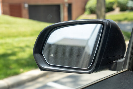 Car side view mirror with blind spot indicator reflecting a residential neighborhood. Concept of road safety, driver assistance, visibility, and modern automotive technology