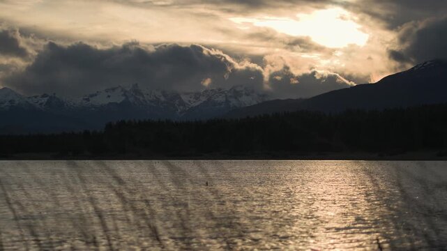 The view of a mountain range with snow-capped peaks seen over a calm lake through foreground grass at sunset.